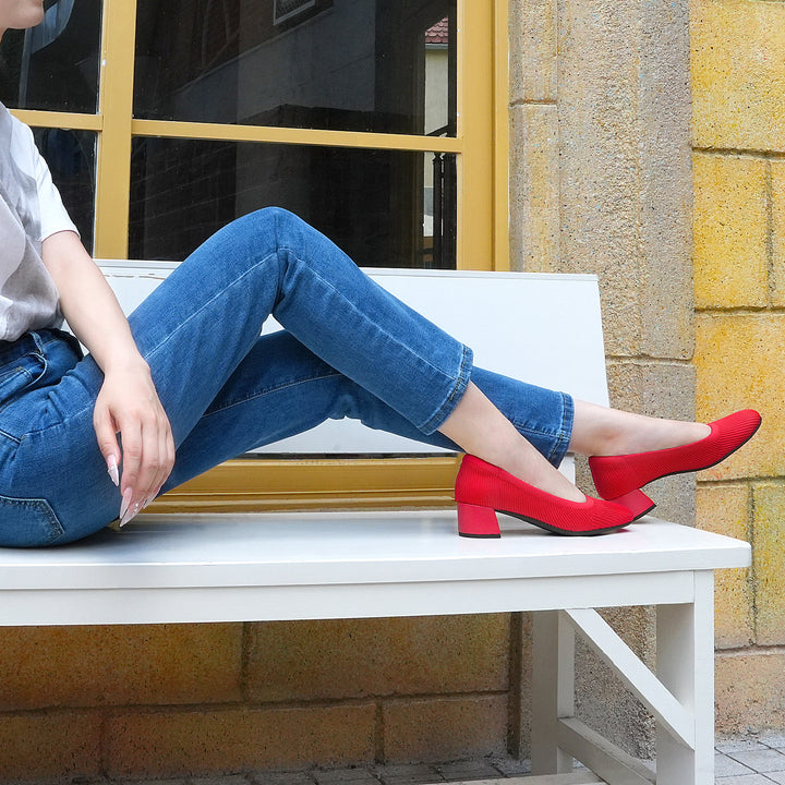 Person sitting on a bench wearing blue jeans and red shoes, with a yellow building in the background.#color_red