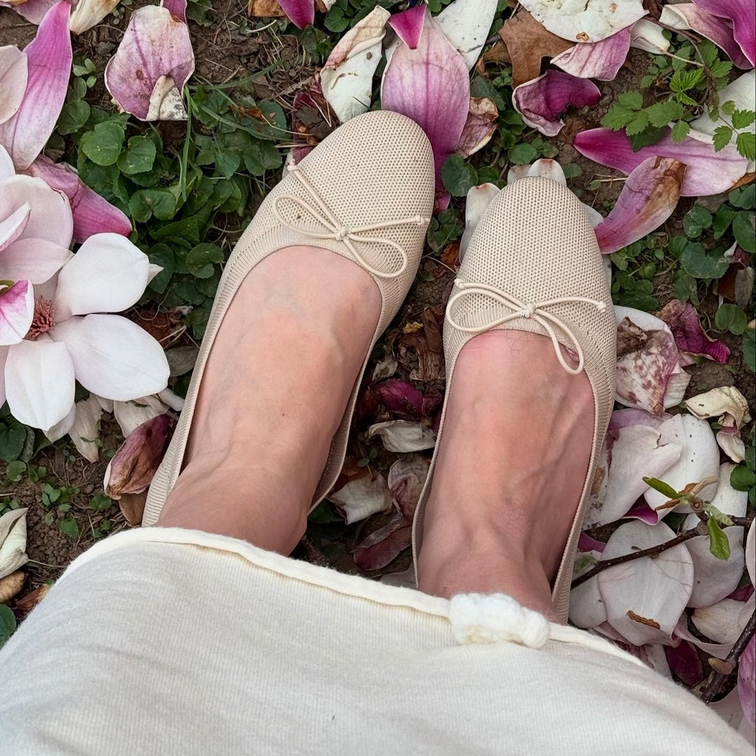 Beige flats with bow details worn on a person's feet, standing on a ground covered with fallen pink and white flowers.#color_beige