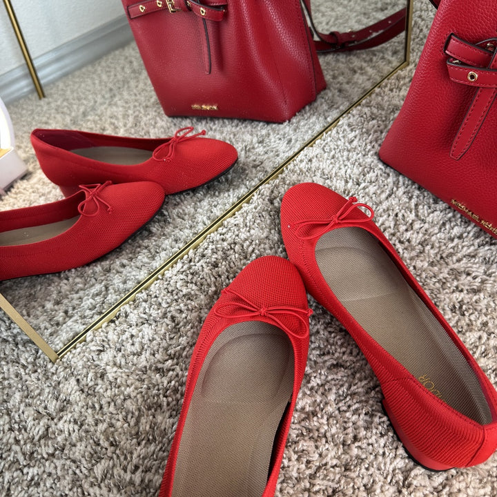 Red shoes with a bow on a carpeted floor next to a red handbag.#color_red