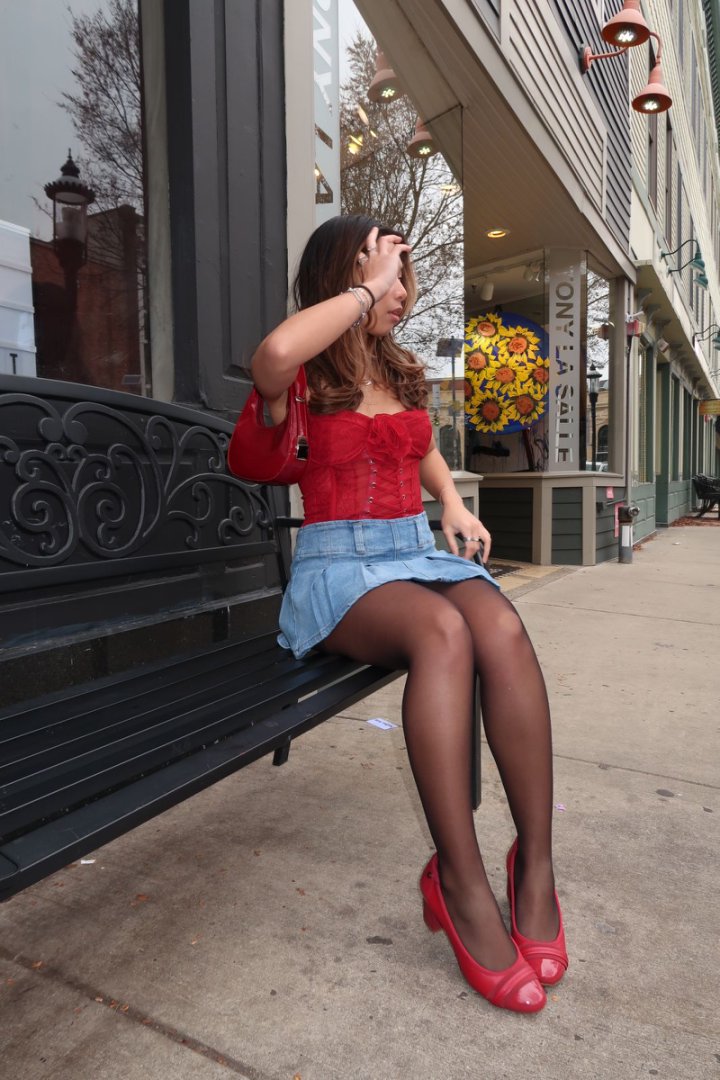 Woman sitting on a bench wearing a red top, denim skirt, and red shoes in an urban setting.#color_red