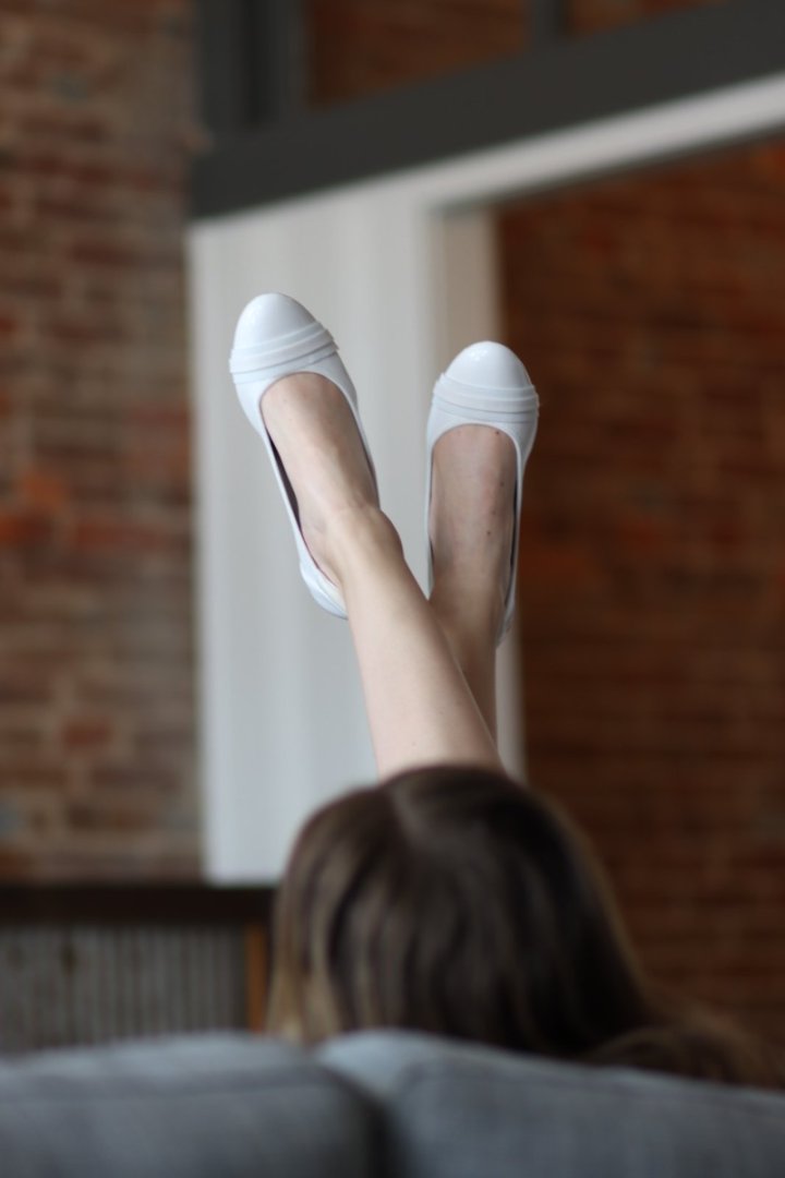 Person wearing white shoes and sheer stockings, sitting on a couch with a brick wall background#color_white