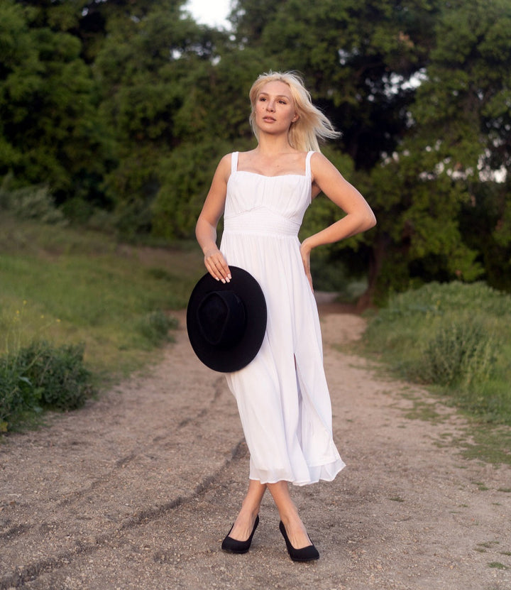 Woman in a white dress holding a black hat on a dirt path with greenery.#color_black1
