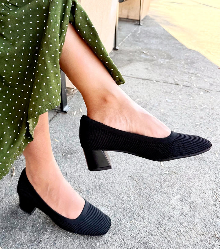 Person wearing black shoes and a green polka dot skirt on a sidewalk.#color_black
