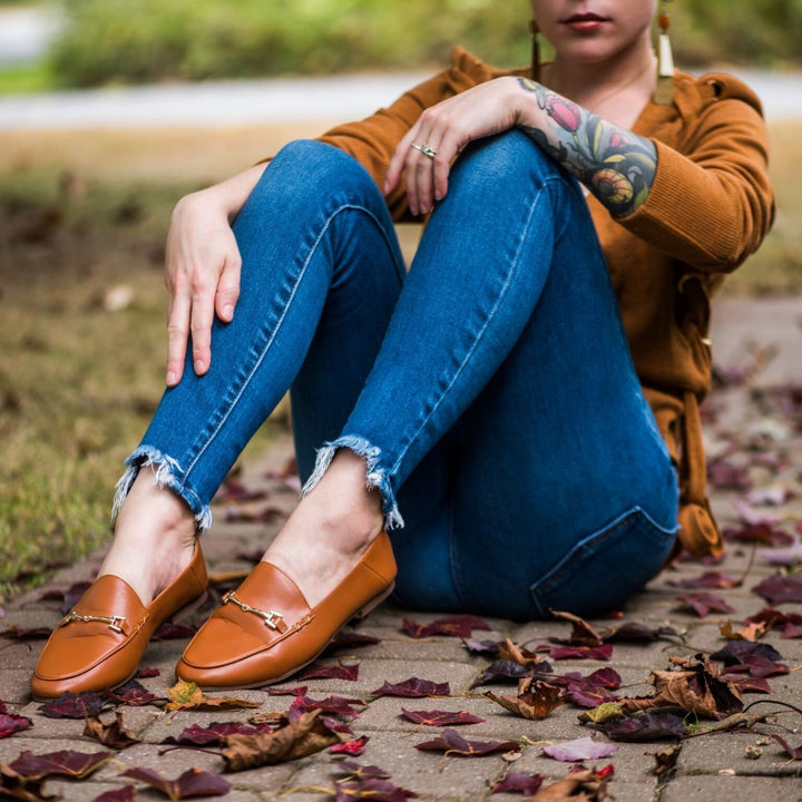 Person wearing brown loafers and blue jeans sitting on a leaf-covered ground.#color_brown