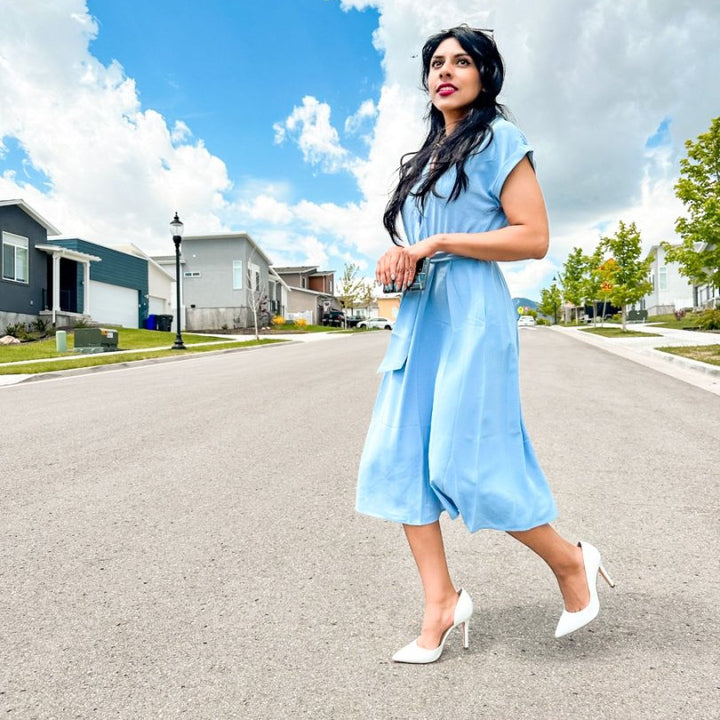 Woman in a light blue dress walking on a street with houses and trees in the background#color_p-white