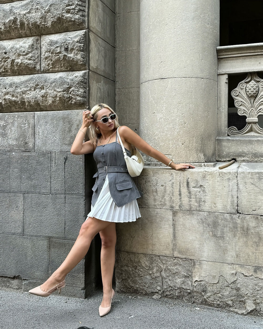 Woman in a gray dress and sunglasses styling slingback kitten heels leaning against a stone wall.#color_nude