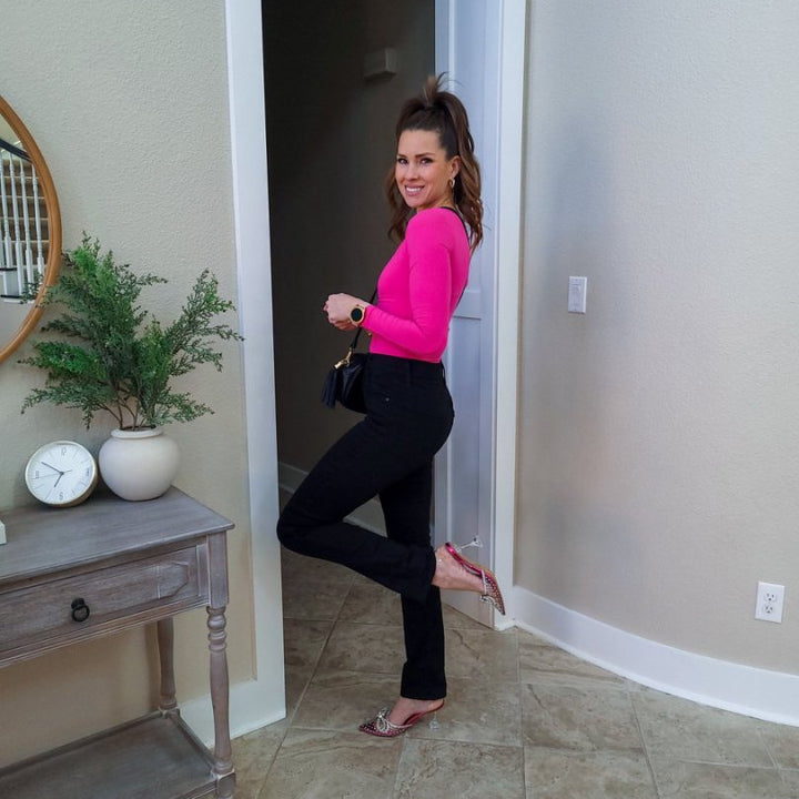 Woman in a pink top and black pants standing in a doorway with a plant and clock on a side table.#color_fuchsia-pink-rhinestone