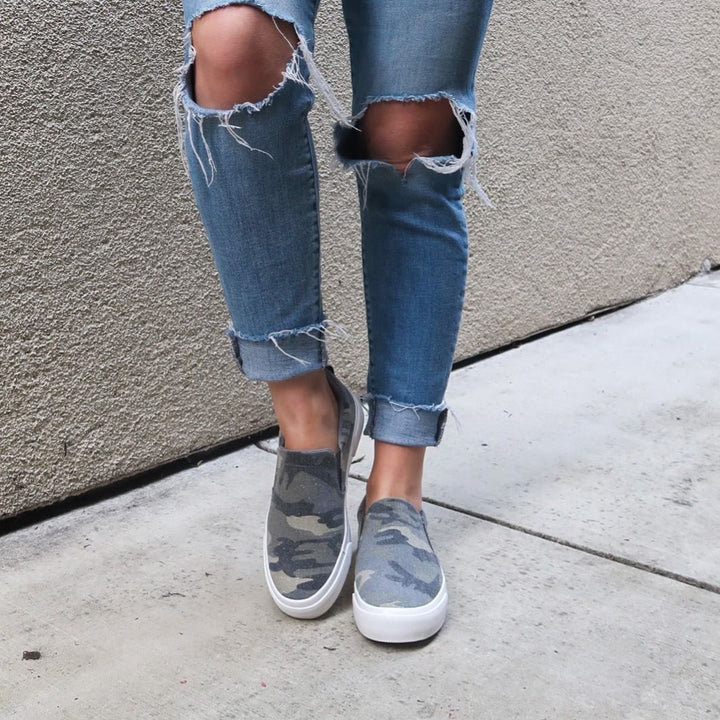 Person wearing ripped jeans and camouflage sneakers against a gray wall.#color_camo