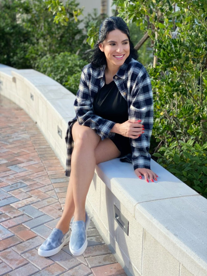 Woman sitting on a stone wall outdoors wearing a plaid shirt and denim shoes.#color_grey-tie-dye
