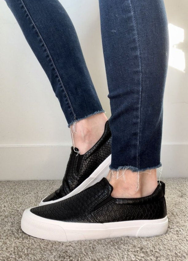 Person wearing black slip-on sneakers with white soles and blue jeans on a neutral background#color_stone-black