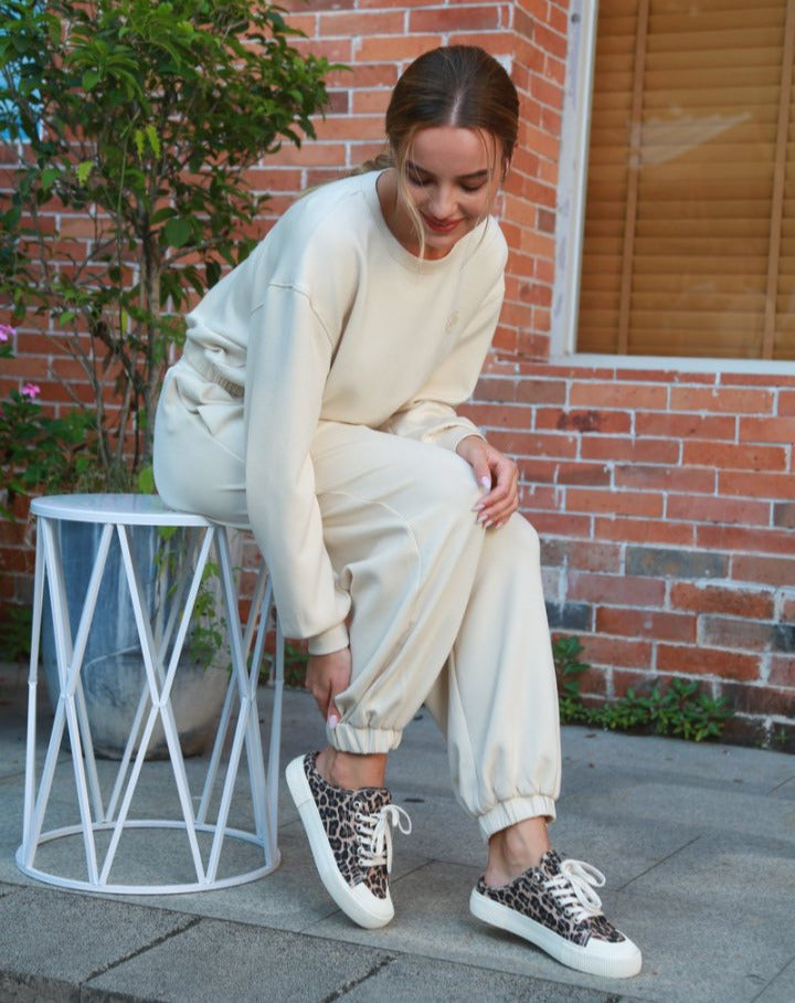 Woman in beige outfit with leopard print sneakers sitting on a white stool against a brick wall.#color_leopard