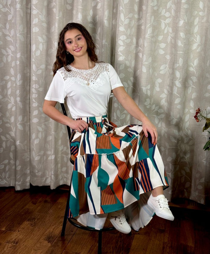 Woman sitting on a chair wearing a white top and colorful patterned skirt against a curtain background#color_white