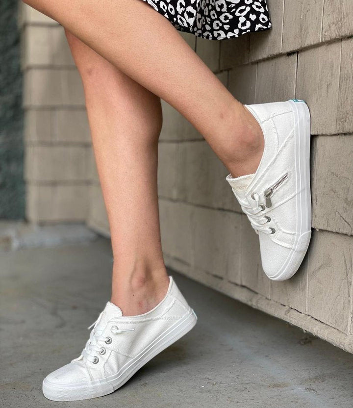 White sneakers worn by a person sitting against a stone wall.#color_white
