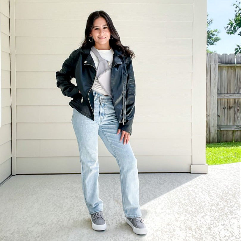 Person wearing a black jacket, white shirt, and light blue jeans standing in front of a white wall.#color_light-grey1