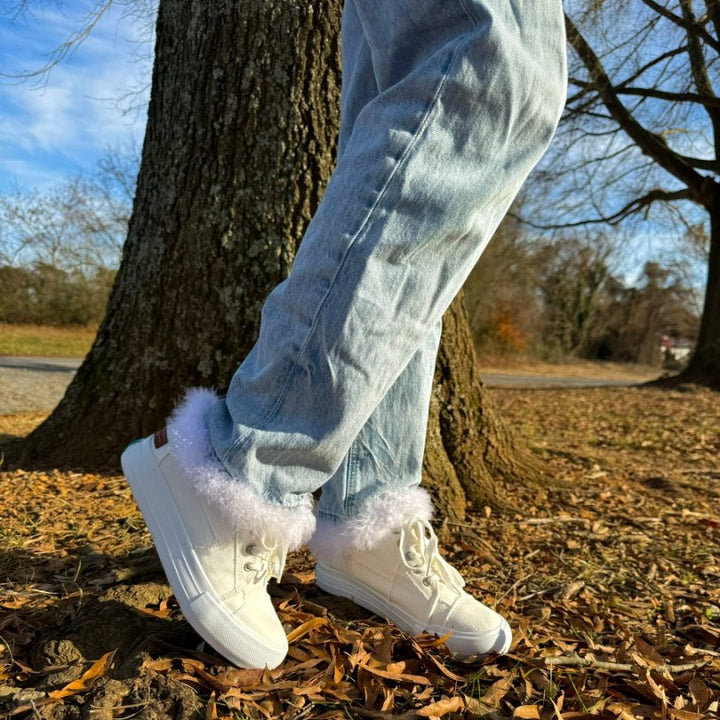 Person wearing white sneakers with fur trim standing on a leaf-covered ground with trees in the background.#color_white1