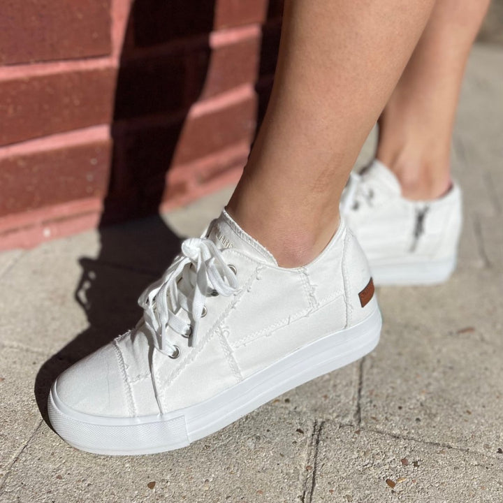 White sneakers worn on a concrete surface with a brick wall in the background#color_white