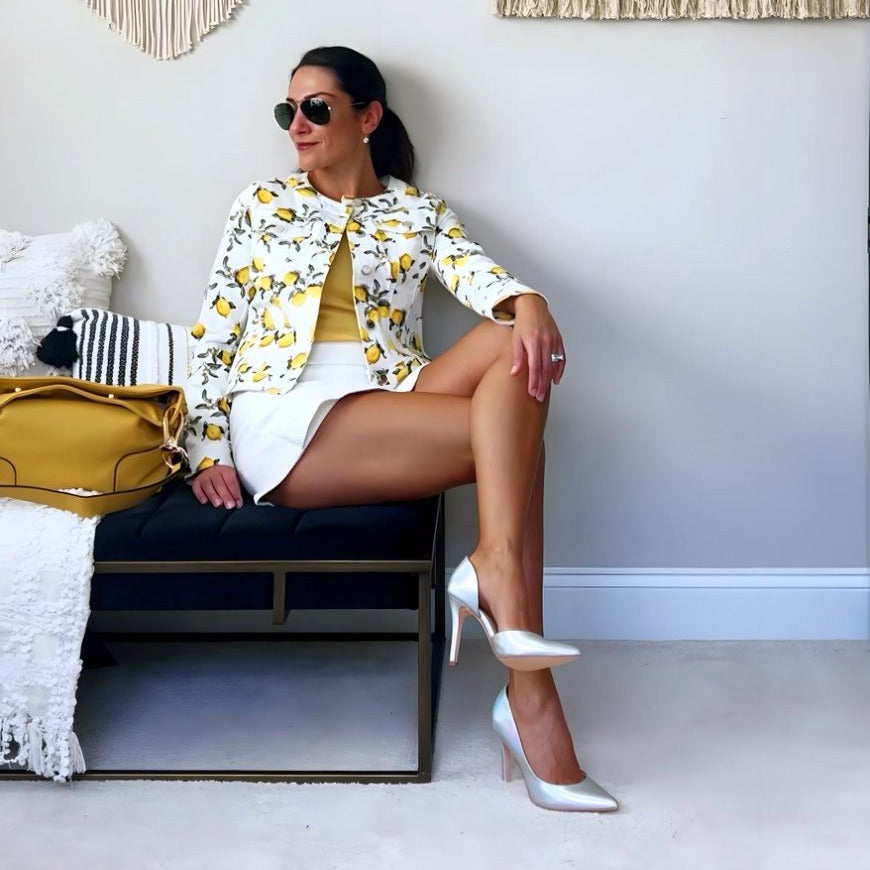 Woman sitting on a bench wearing a patterned jacket and white high heels in a room with decorative wall art.#color_light-blue