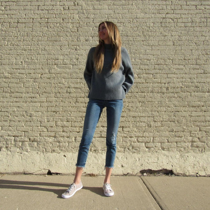 Woman in a gray sweater and blue jeans standing against a brick wall.#color_light-grey