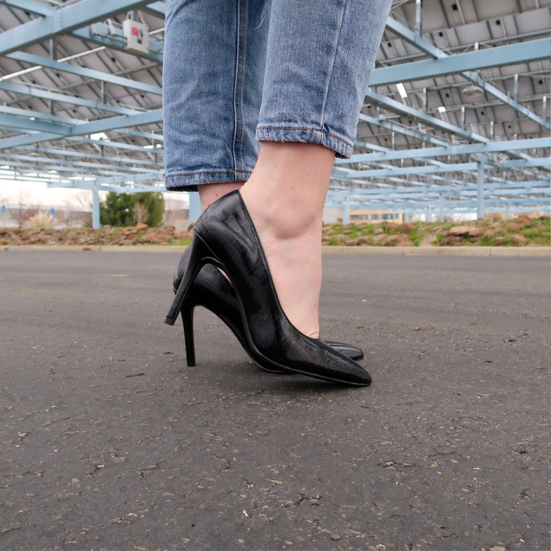 Person wearing black high-heeled shoes and blue jeans standing on a paved surface with an industrial background.#color_p-black