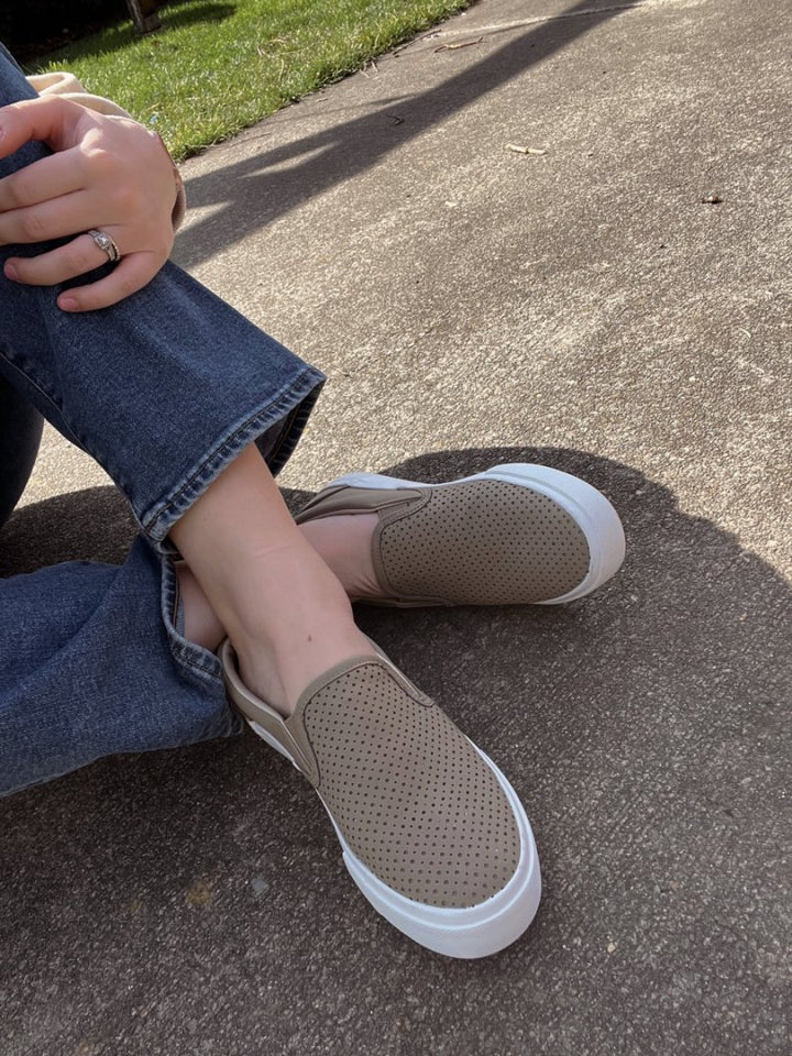 Person wearing brown slip-on shoes with white soles on a concrete surface.#color_taupe