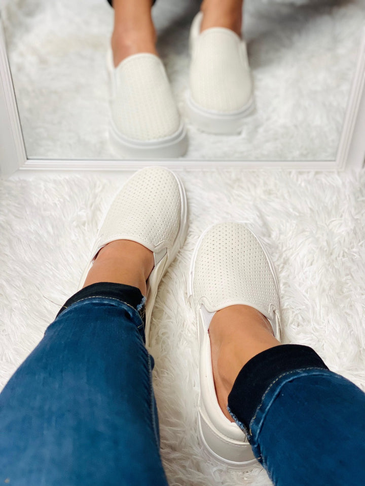 White slip-on shoes worn with blue jeans on a white floor.#color_white