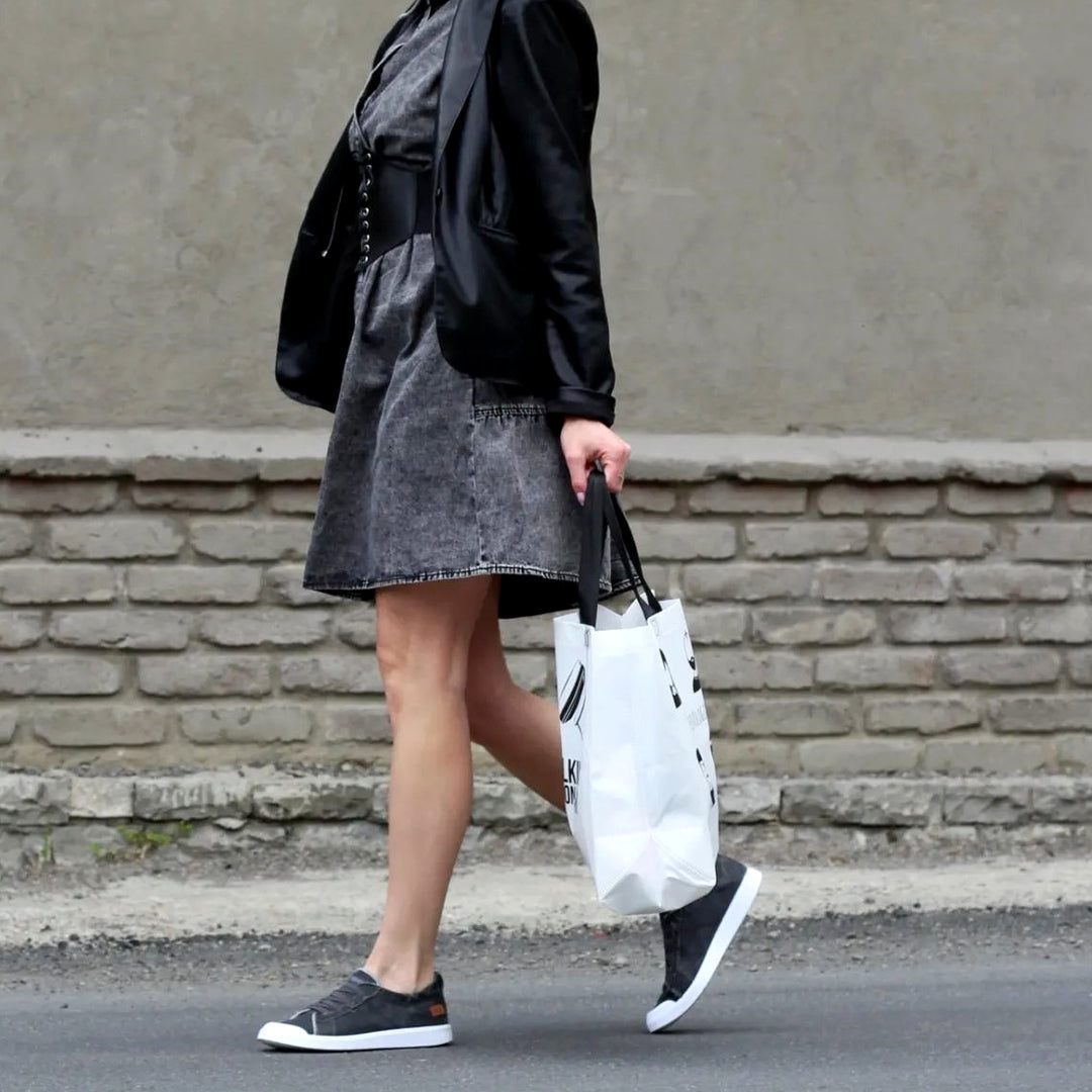 Person walking on a street holding a white bag, wearing a black jacket and denim skirt.#color_dark-grey
