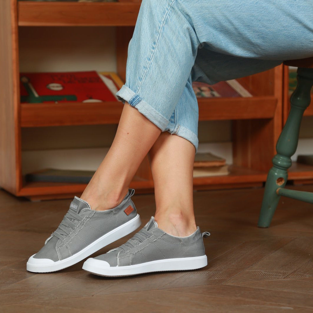 Person wearing gray sneakers on a wooden floor with a bookshelf in the background#color_light-grey